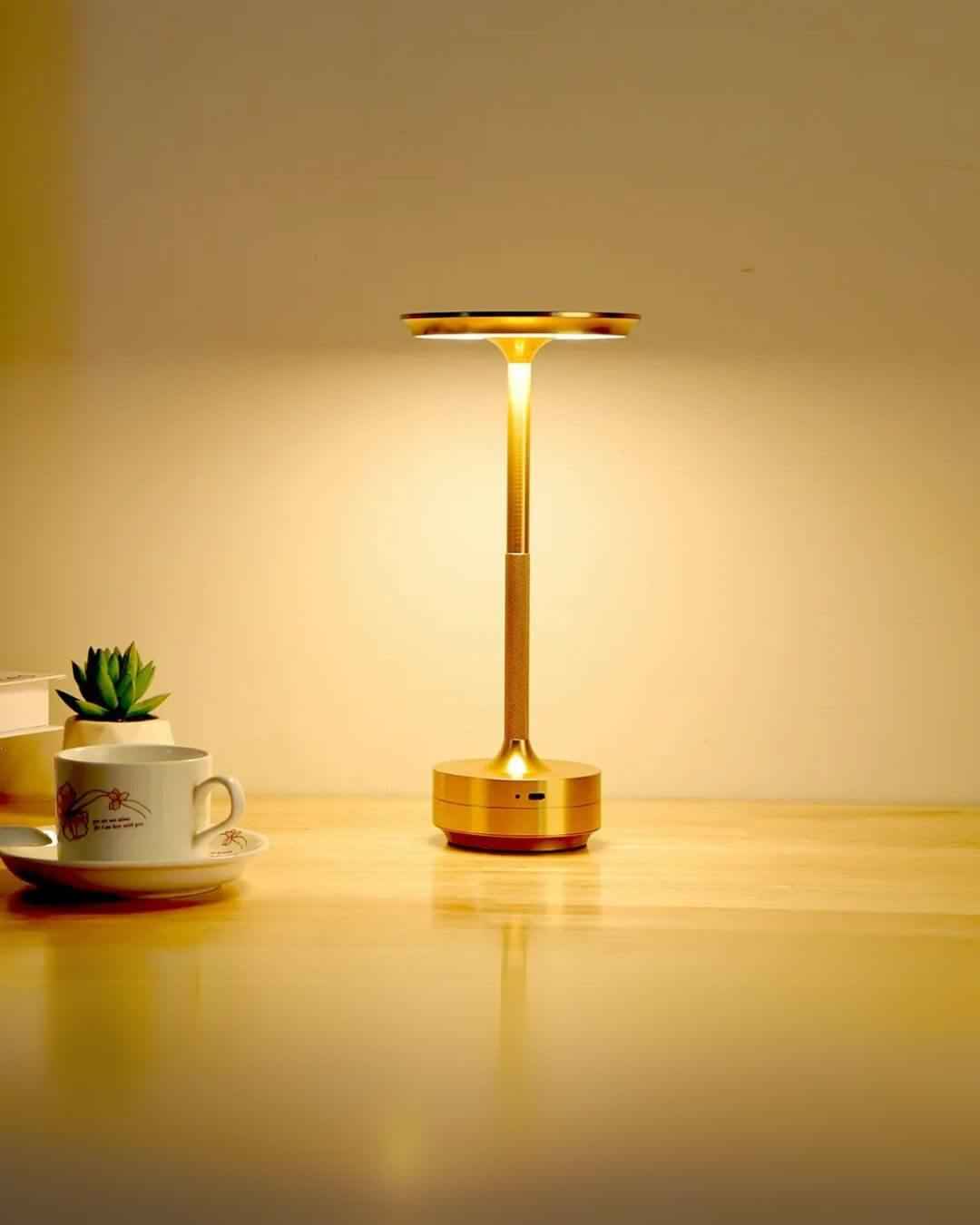 LumiHikari Base Lamp with brushed metal finish providing warm ambient light on wooden desk next to a coffee cup and small plant.