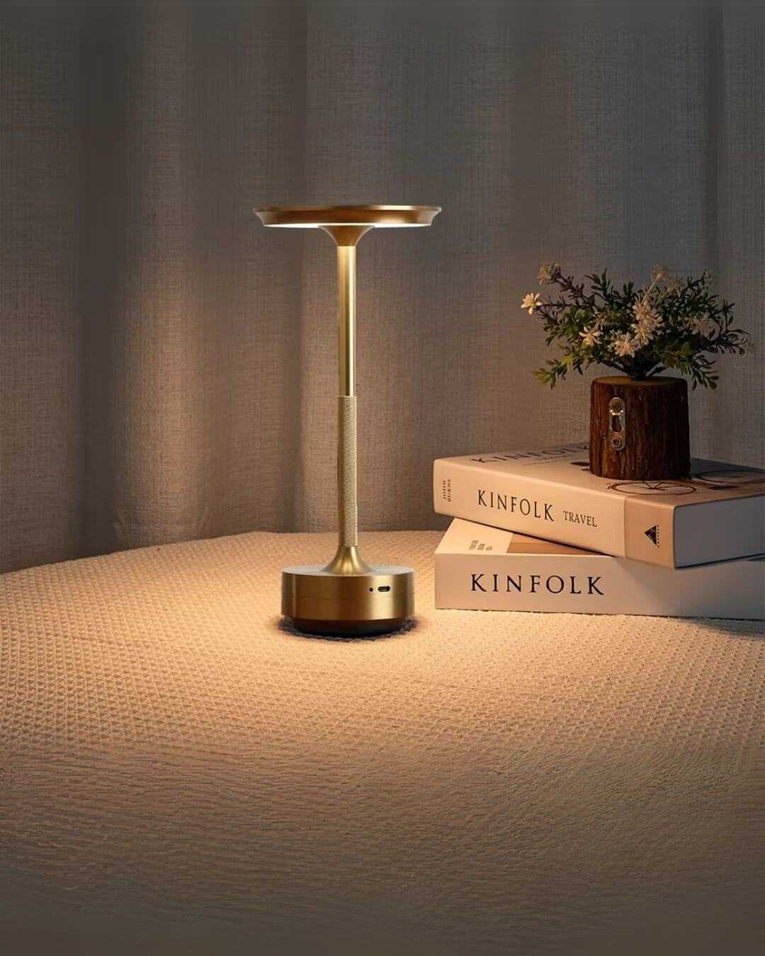 Modern LumiHikari Base Lamp with brushed metal finish providing warm ambient light on a textured surface next to stacked books and small plant.